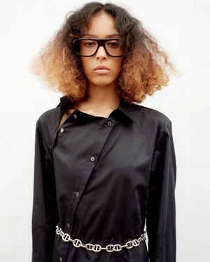 Model Prinnie Stott wearing a black button-down shirt, oversized dark glasses, and a silver chain belt against a white studio background.