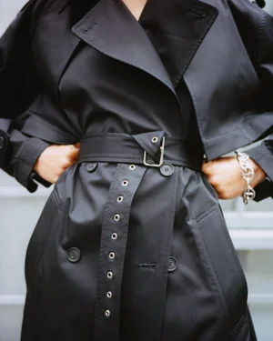Model wearing a black belted trench coat with metal eyelets and a silver chain bracelet for Eudon Choi Resort 25