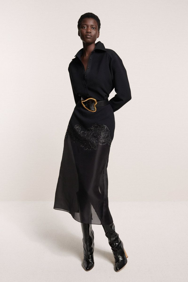 Full-length shot of a Black woman in a black collared dress with a heart-shaped belt buckle and shiny black boots against a white backdrop.
