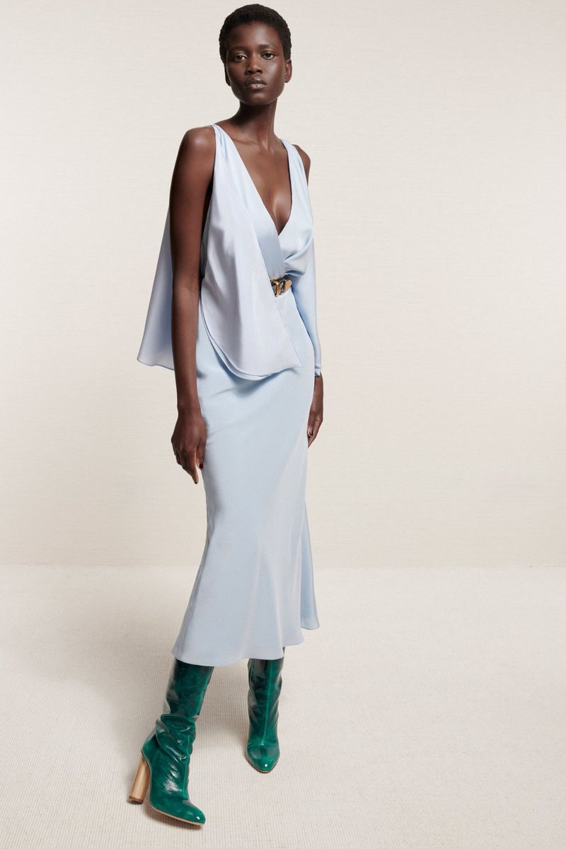 Full-length shot of a slender African woman in a light blue dress and green boots against a neutral background in a fashion editorial.