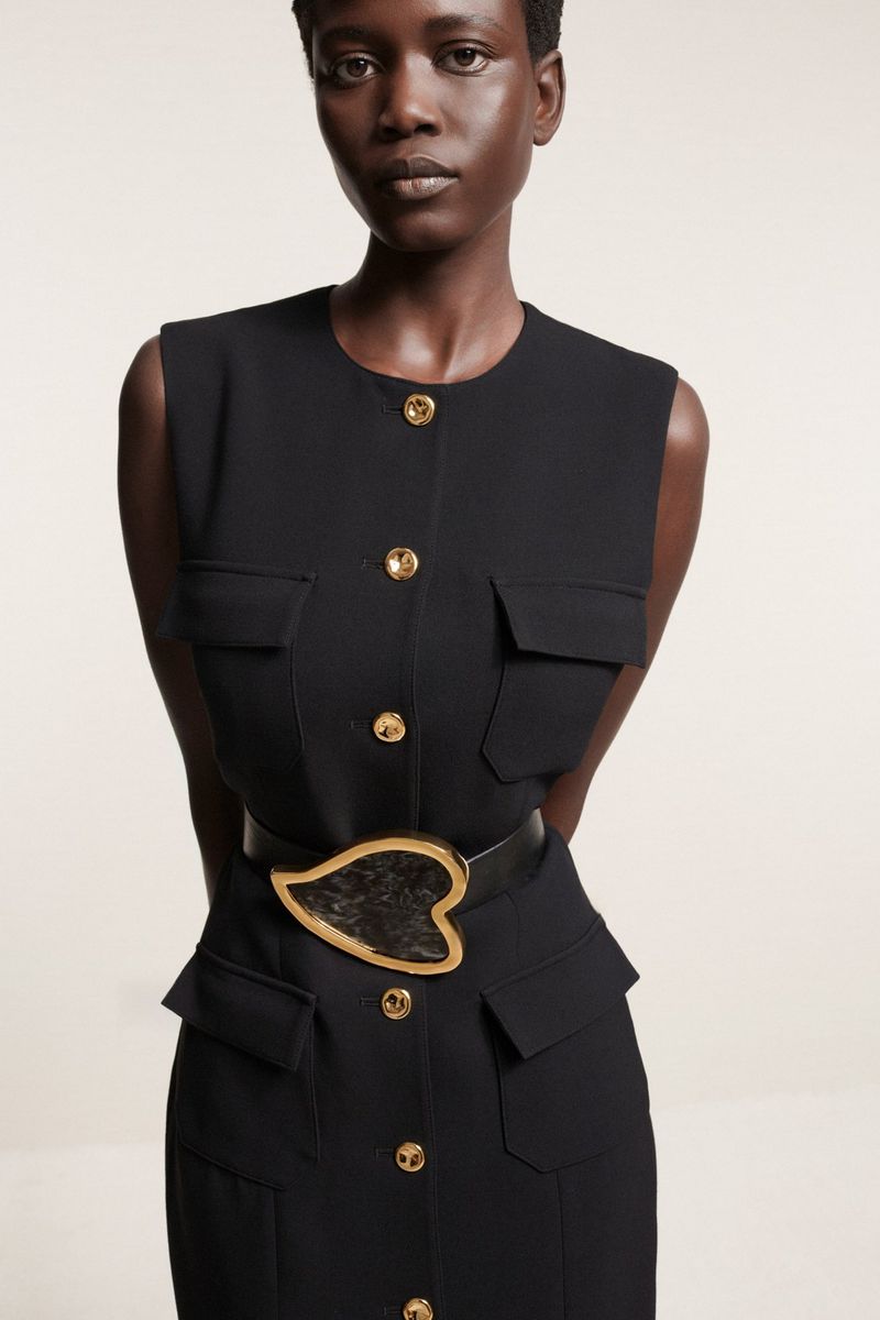 An elegant Black woman poses indoors wearing a black sleeveless dress with gold buttons, cinched at the waist with a statement belt.