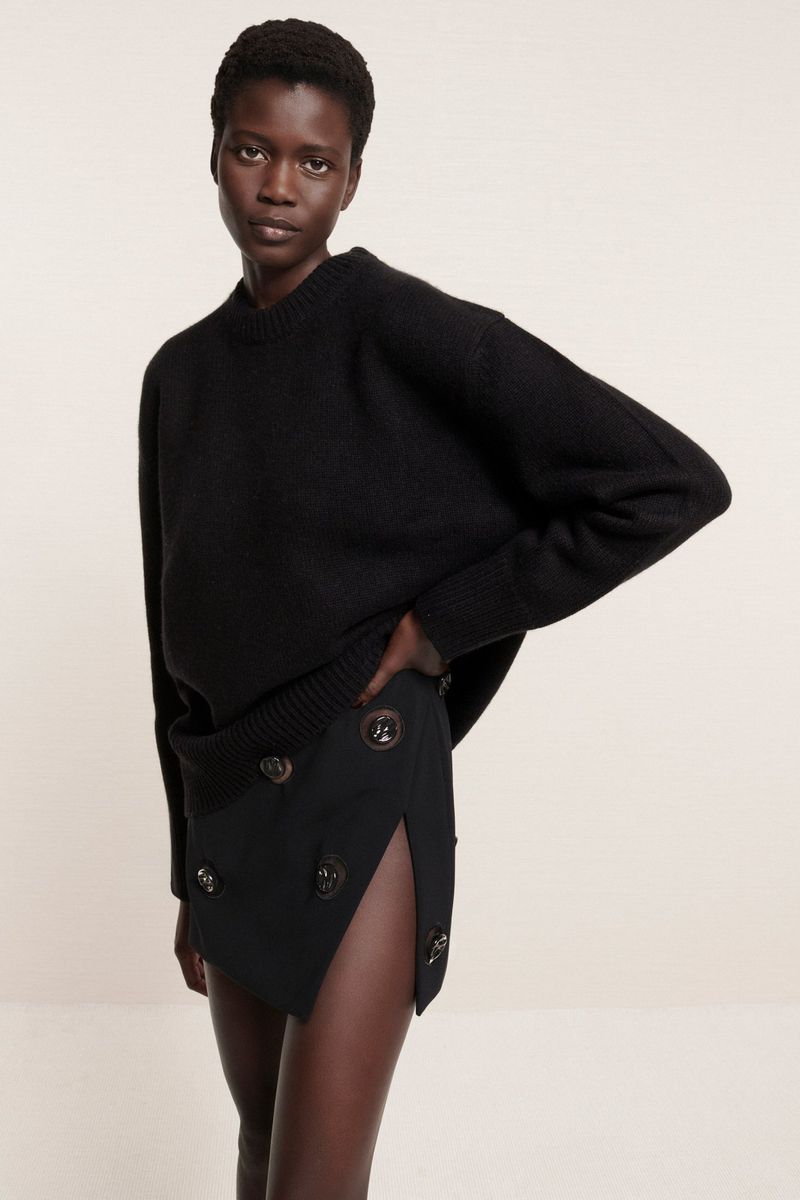 Portrait of a Black woman in a black sweater and black skirt with buttons posing in front of a neutral background, fashion shot.