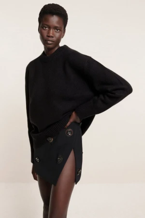 Portrait of a Black woman in a black sweater and black skirt with buttons posing in front of a neutral background, fashion shot.