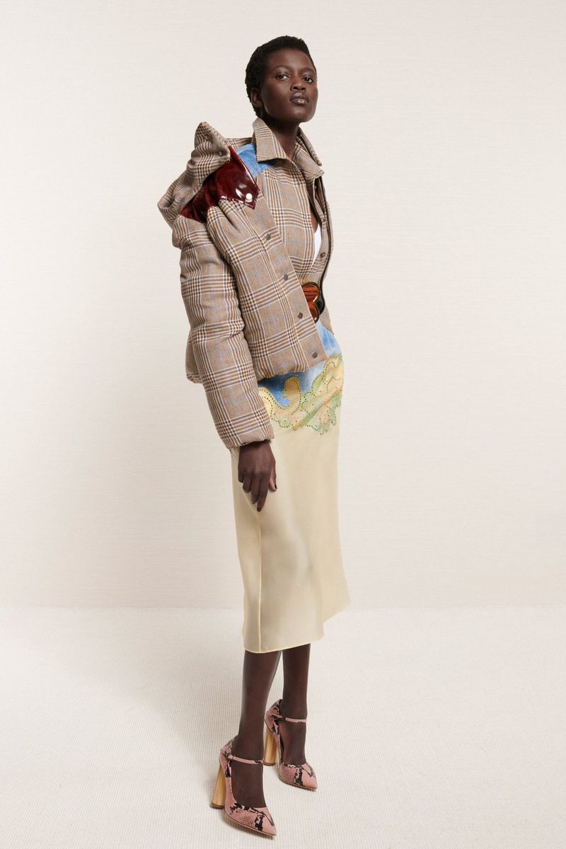 A dark-skinned female model poses in a studio wearing a plaid puffer jacket with a patterned top and cream-colored skirt, accessorized with patterned heels.