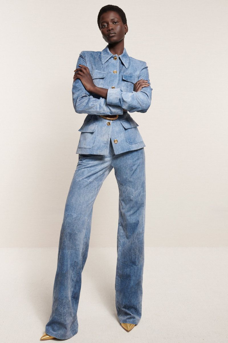 A full-length shot of a Black woman in a light blue denim jacket and bell-bottom pants, posing against a neutral backdrop in a fashion editorial.
