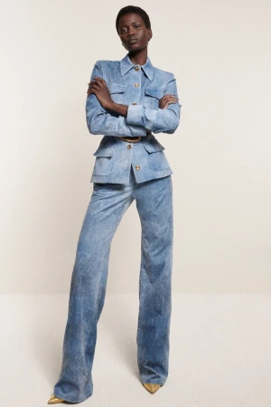 A full-length shot of a Black woman in a light blue denim jacket and bell-bottom pants, posing against a neutral backdrop in a fashion editorial.