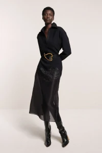 Model in black shirt dress with heart-shaped belt and tall boots standing against a neutral background
