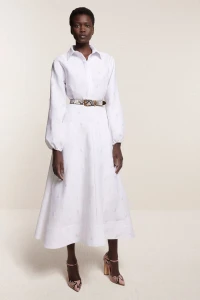 Model wearing a white midi shirt dress with a patterned belt against a cream backdrop for Brandon Maxwell Resort 2025 lookbook.