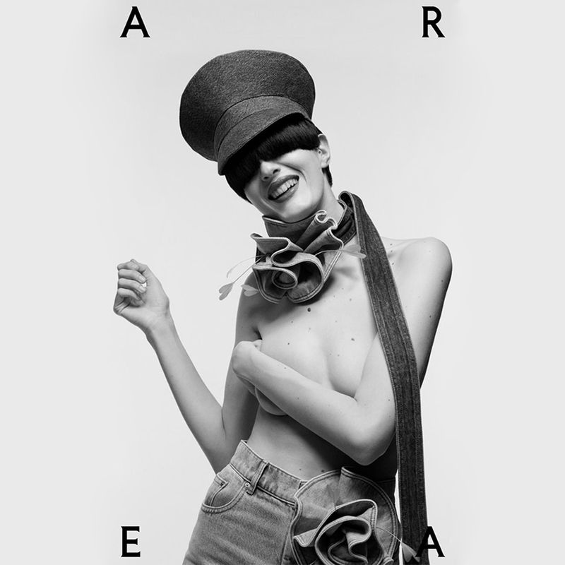 Black and white studio portrait of a smiling topless model with denim jeans and flower embellishments, wearing a dark hat that covers her eyes.
