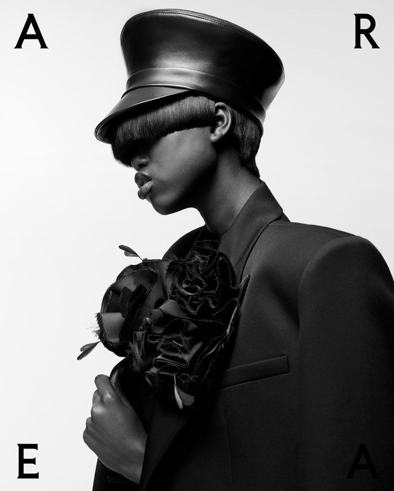 Monochrome portrait of androgynous person in peaked cap and suit jacket with fabric flower against a white backdrop, with letters.