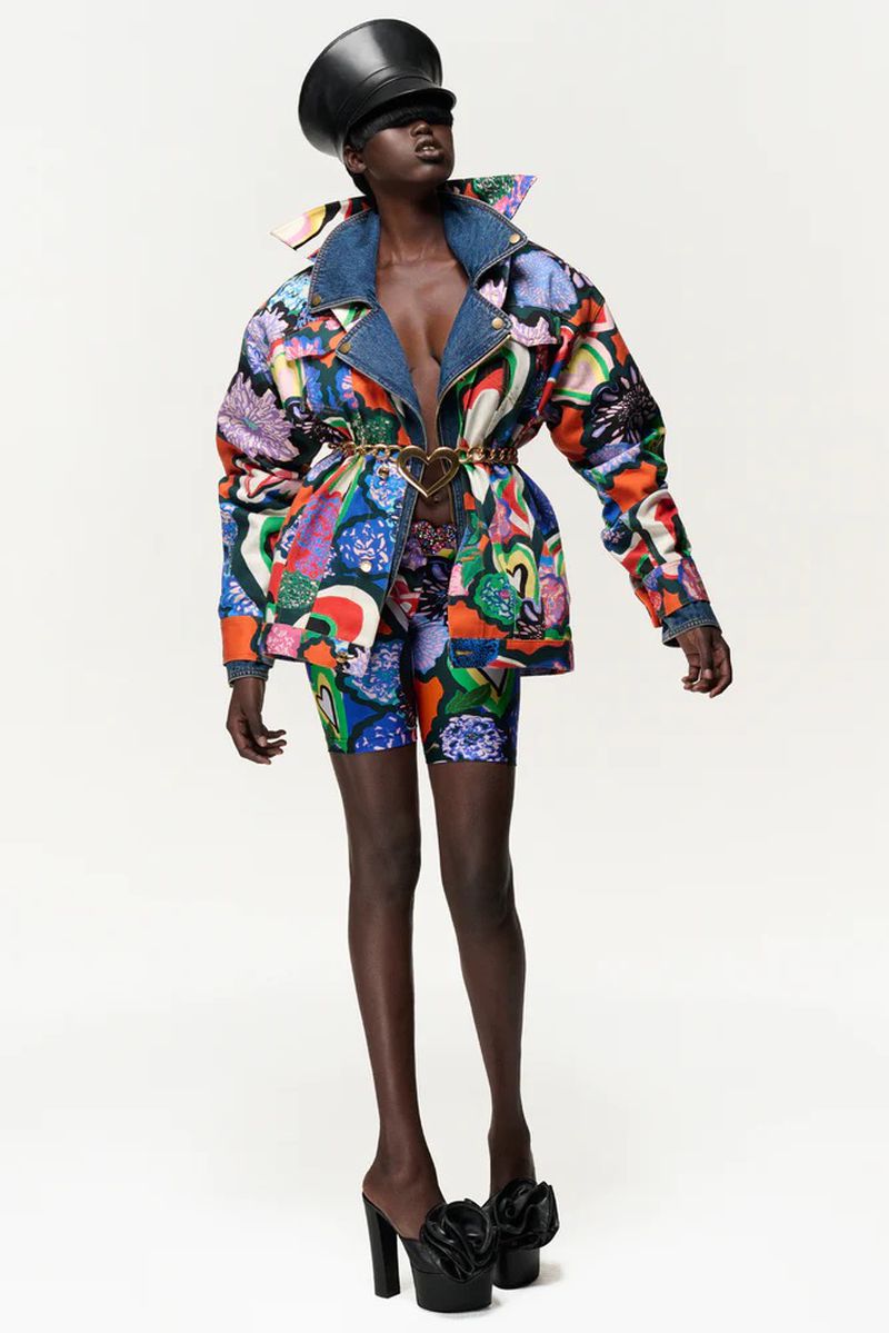 A woman models a colorful patterned denim jacket and shorts with platform shoes in a studio setting, wearing a black leather cap.