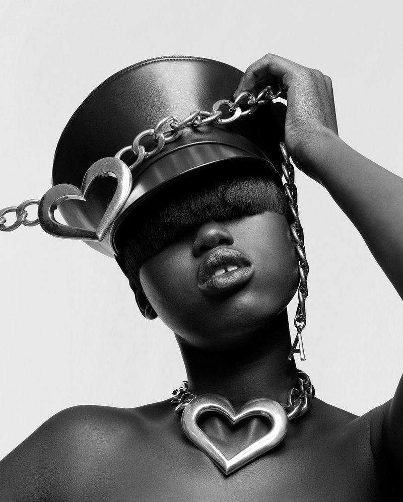 Black and white portrait of an adult female with dark skin wearing a hat, chain, and heart necklace, captured in a studio setting.