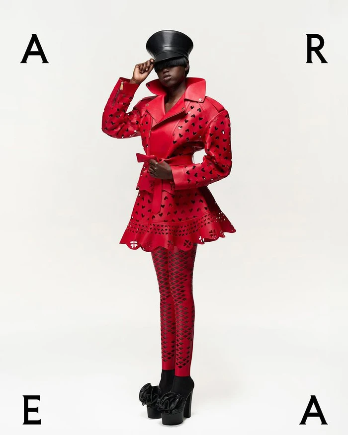 Model in red cutout jacket and matching patterned tights wearing a black cap against a plain white background