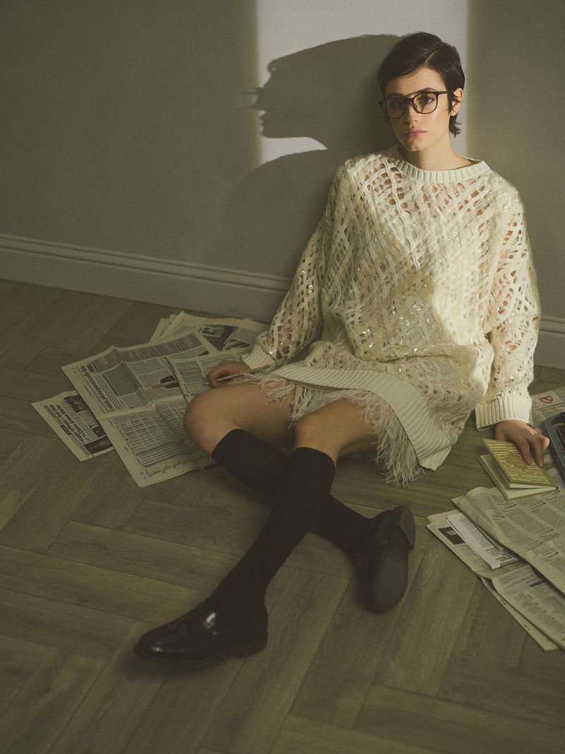 Young woman with short hair, glasses, and a sweater dress sits on the floor surrounded by newspapers in a softly lit indoor space.