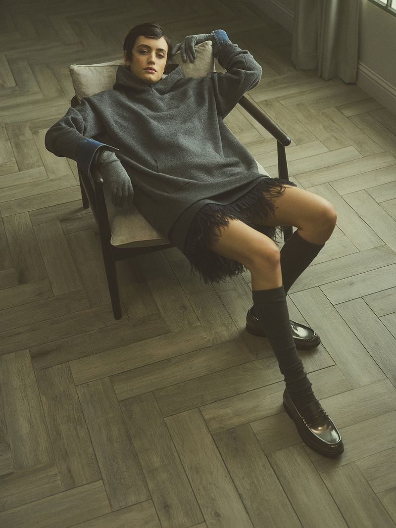 Indoor fashion shot of a woman in a grey sweater dress, knee socks, and loafers sitting in a chair with parquet flooring.