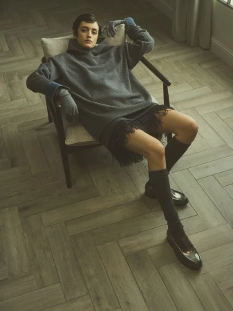 Indoor fashion shot of a woman in a grey sweater dress, knee socks, and loafers sitting in a chair with parquet flooring.