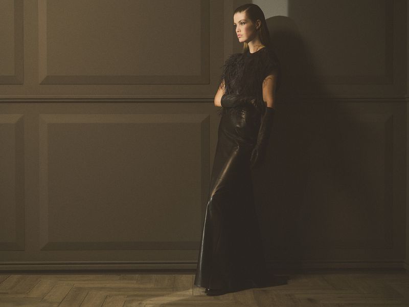 Elegant woman in long black dress and gloves stands against a paneled wall with soft light casting shadows, creating a sophisticated atmosphere.
