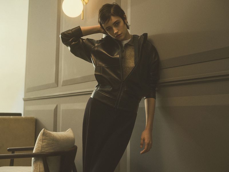 A young woman with short dark hair poses in a black leather jacket and pants against a grey wall, under a warm-toned wall sconce light.