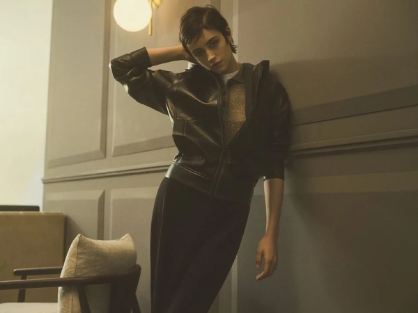 A young woman with short dark hair poses in a black leather jacket and pants against a grey wall, under a warm-toned wall sconce light.