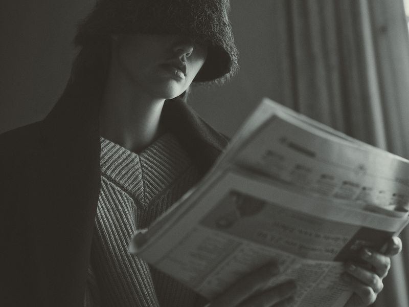 Monochrome portrait of a person in a fur hat partially obscuring their face, reading a newspaper indoors, wearing a striped sweater.