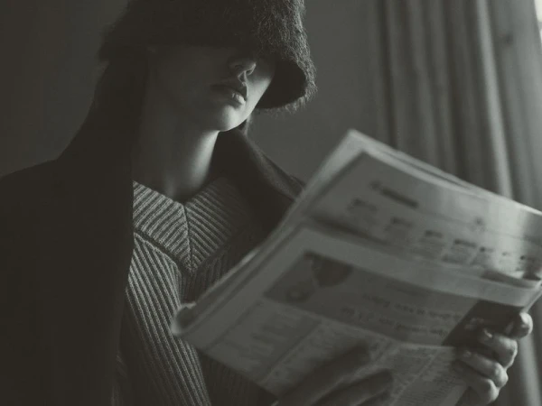 Monochrome portrait of a person in a fur hat partially obscuring their face, reading a newspaper indoors, wearing a striped sweater.