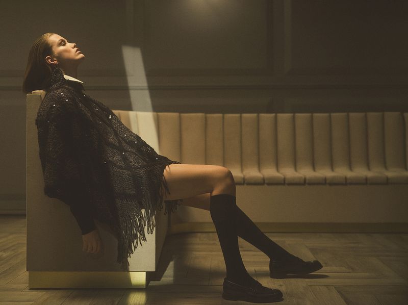 Adult female model with long legs poses in black clothing and knee-high socks on a neutral platform in a studio with parquet flooring.