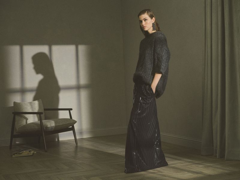 A woman in a gray sweater and black skirt poses in a dimly lit room, with a modern chair and a shadow on the wall.