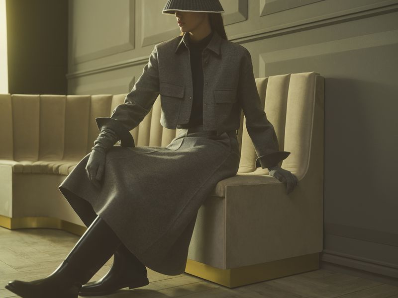 Elegant woman in grey skirt suit, gloves and high boots poses indoors on stylish bench seating in a fashion photography setting.