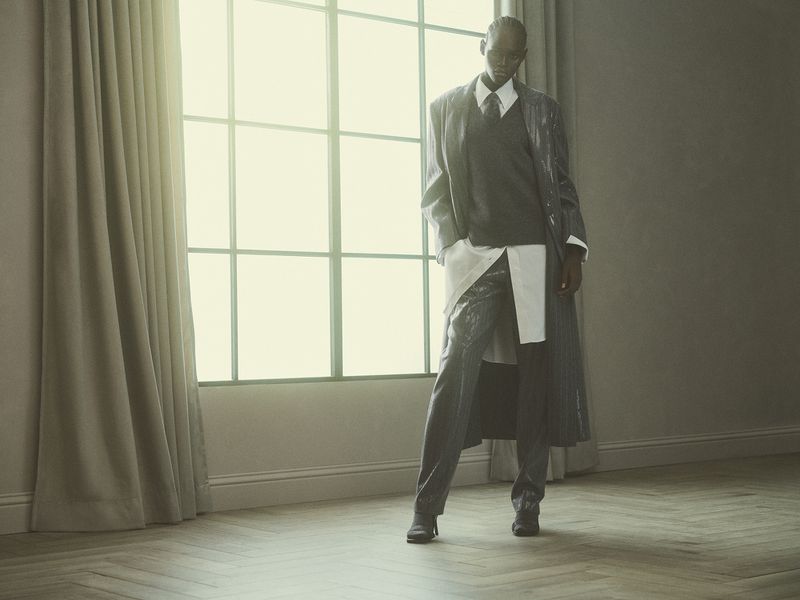 Model Ageer Ayuen poses in a layered grey coat, sweater vest, collared shirt, and trousers by a large window for Brunello Cucinelli.
