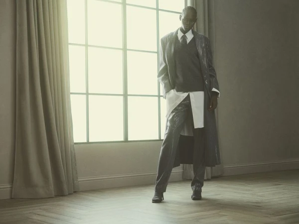 Model Ageer Ayuen poses in a layered grey coat, sweater vest, collared shirt, and trousers by a large window for Brunello Cucinelli.