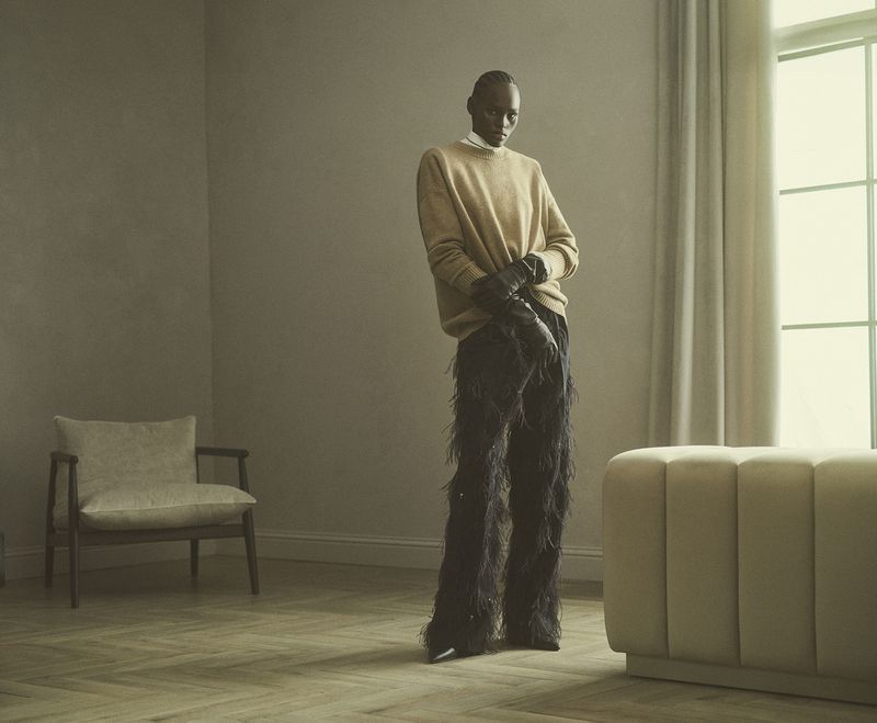 Full-length studio shot of a Black model wearing a tan sweater and black feather pants, standing on a herringbone wood floor.