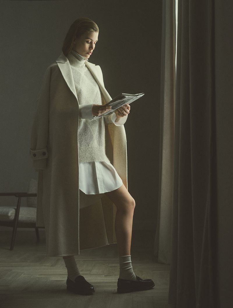 Indoor shot of a young fair-skinned woman reading a newspaper. She's wearing a turtleneck, sweater, skirt, coat, socks, and loafers.