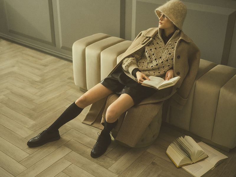 A woman is sitting indoors reading a book wearing a beige knit sweater, black shorts, black knee socks, and black leather shoes.
