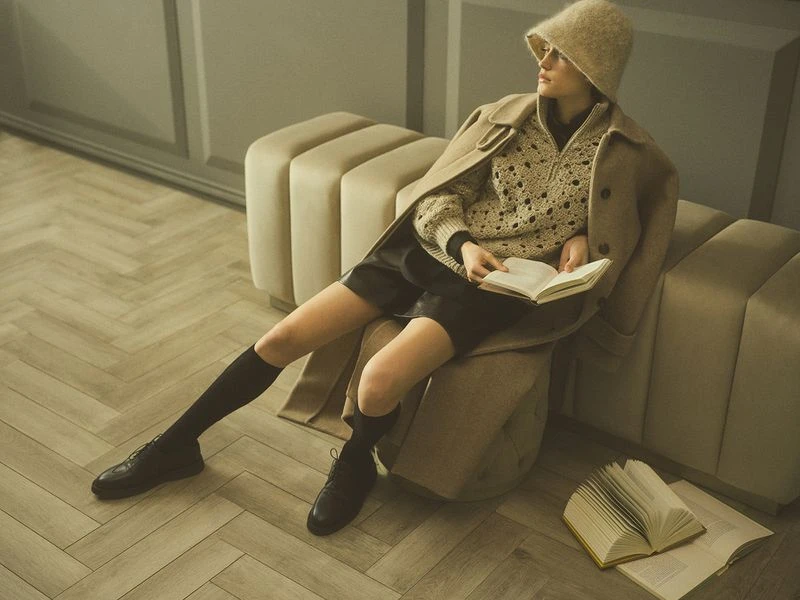 A woman is sitting indoors reading a book wearing a beige knit sweater, black shorts, black knee socks, and black leather shoes.