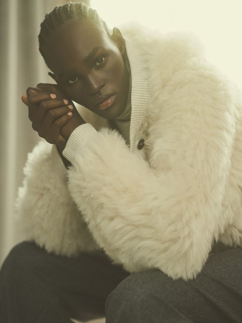 Portrait of a dark-skinned adult male wearing a cream-colored, shearling-style coat with his hands clasped, set against a soft, light background.