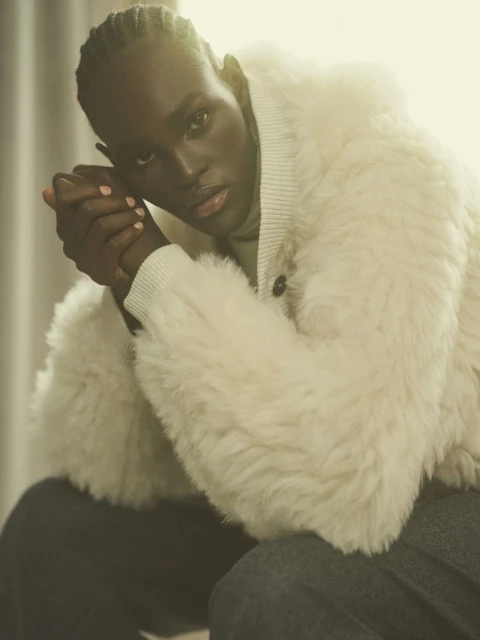 Portrait of a dark-skinned adult male wearing a cream-colored, shearling-style coat with his hands clasped, set against a soft, light background.