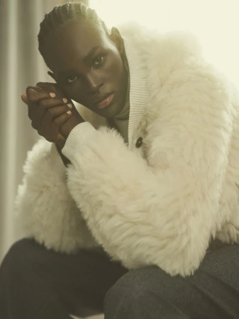 Portrait of a dark-skinned adult male wearing a cream-colored, shearling-style coat with his hands clasped, set against a soft, light background.