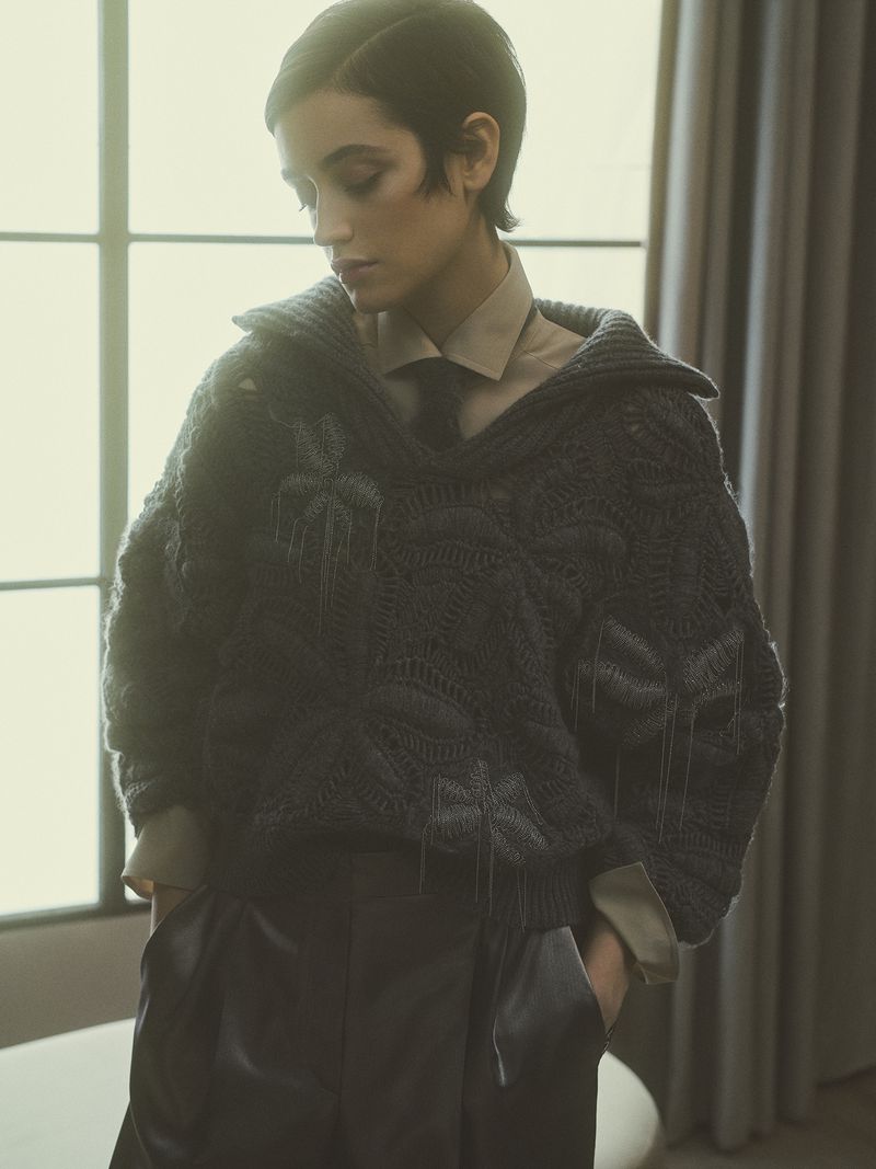 A fashionable woman with short dark hair poses indoors wearing a chunky knit sweater, collared shirt, and stylish trousers, creating a stylish look.