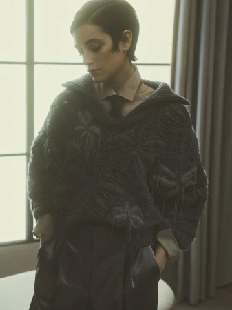 A fashionable woman with short dark hair poses indoors wearing a chunky knit sweater, collared shirt, and stylish trousers, creating a stylish look.