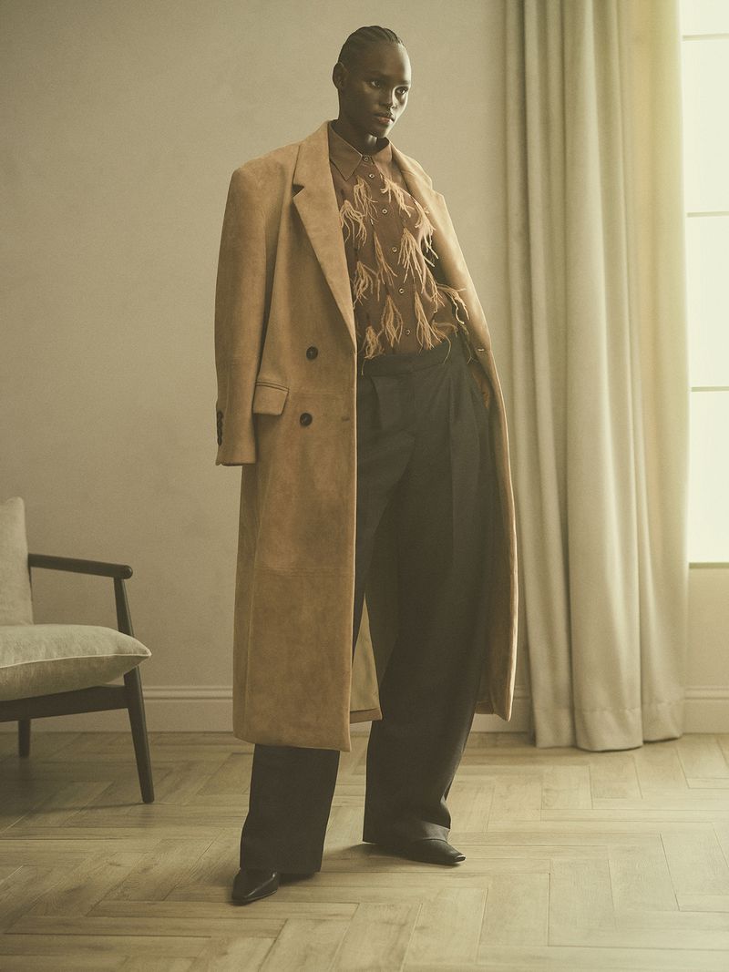 An androgynous adult stands indoors with a tan coat, patterned button-down shirt, and black wide-leg pants, posing in a fashion shot.