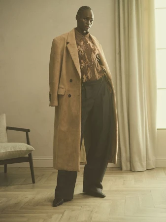 An androgynous adult stands indoors with a tan coat, patterned button-down shirt, and black wide-leg pants, posing in a fashion shot.