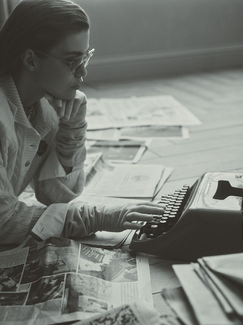 Woman in eyeglasses types on a vintage typewriter, surrounded by newspapers on the floor in a grey-toned indoor setting.
