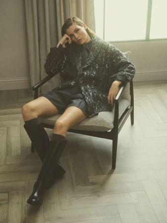 Woman sits in chair wearing a black sequin jacket, shorts, and boots in a neutral-toned interior with herringbone floors.