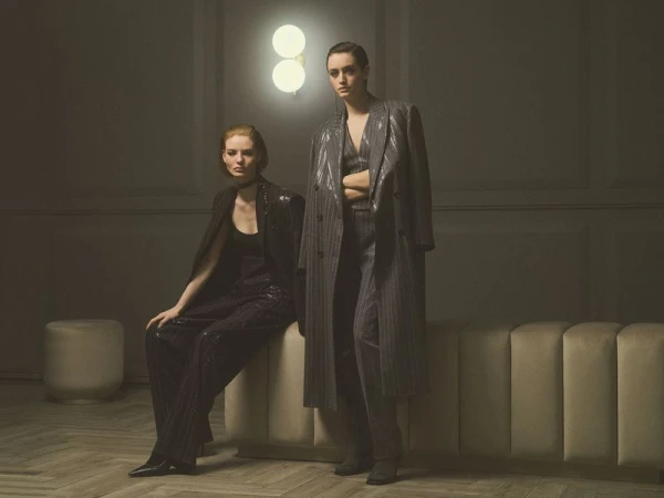 Two women posing in tailored suits and an oversized pinstripe coat beside a glowing wall sconce in a dimly lit room.
