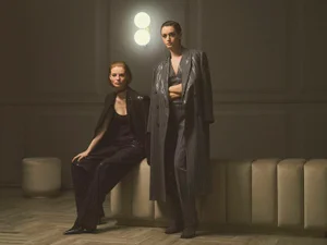 Two women posing in tailored suits and an oversized pinstripe coat beside a glowing wall sconce in a dimly lit room.