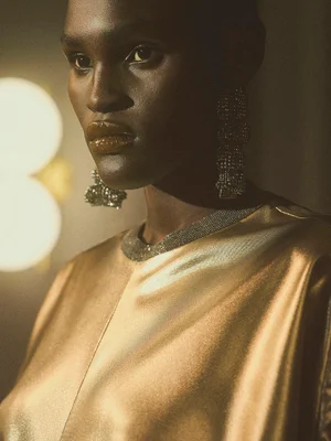 Close-up portrait of a model wearing a metallic gold top and ornate crystal earrings in a warm, softly lit interior setting.