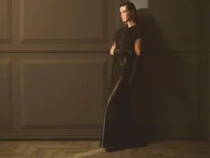 Model wearing a black feathered top and leather skirt standing against a paneled wall in a fashion portrait.