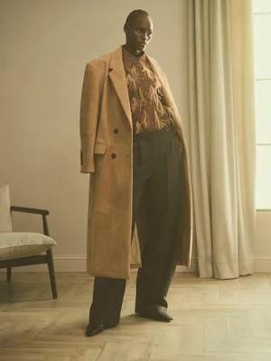 Model wearing a long brown camel coat and dark trousers standing in a bright room with a wooden floor.