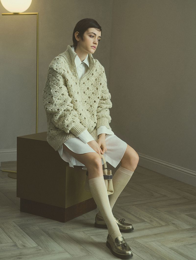 Model seated on a dark cube wearing a cream textured cardigan and white shirt with tall socks and loafers