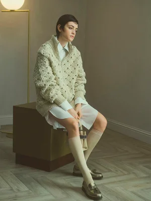 Model seated on a dark cube wearing a cream textured cardigan and white shirt with tall socks and loafers